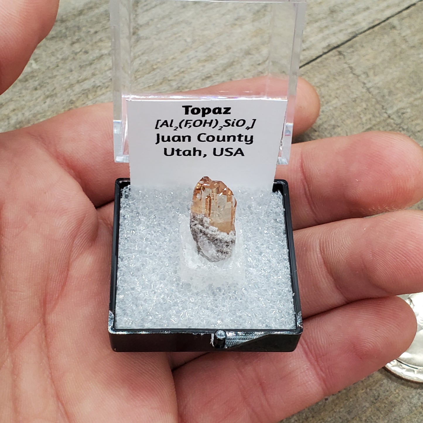 Peach colored topaz crystal in box with formula and origin label.
