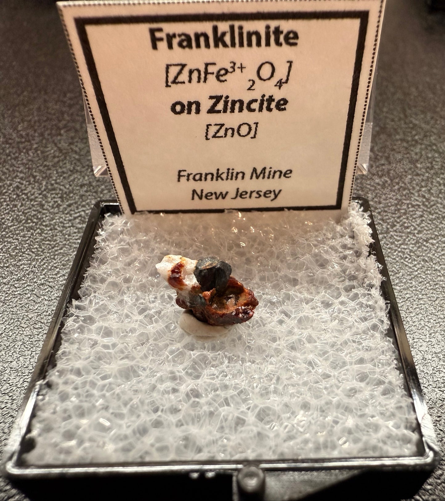 Mineral sample of franklinite on a display case with a label.