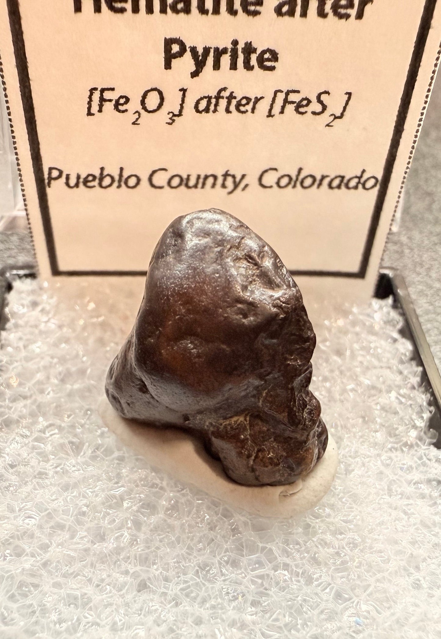 Mineral sample of Hematite after Pyrite on a gray background with descriptive label.
