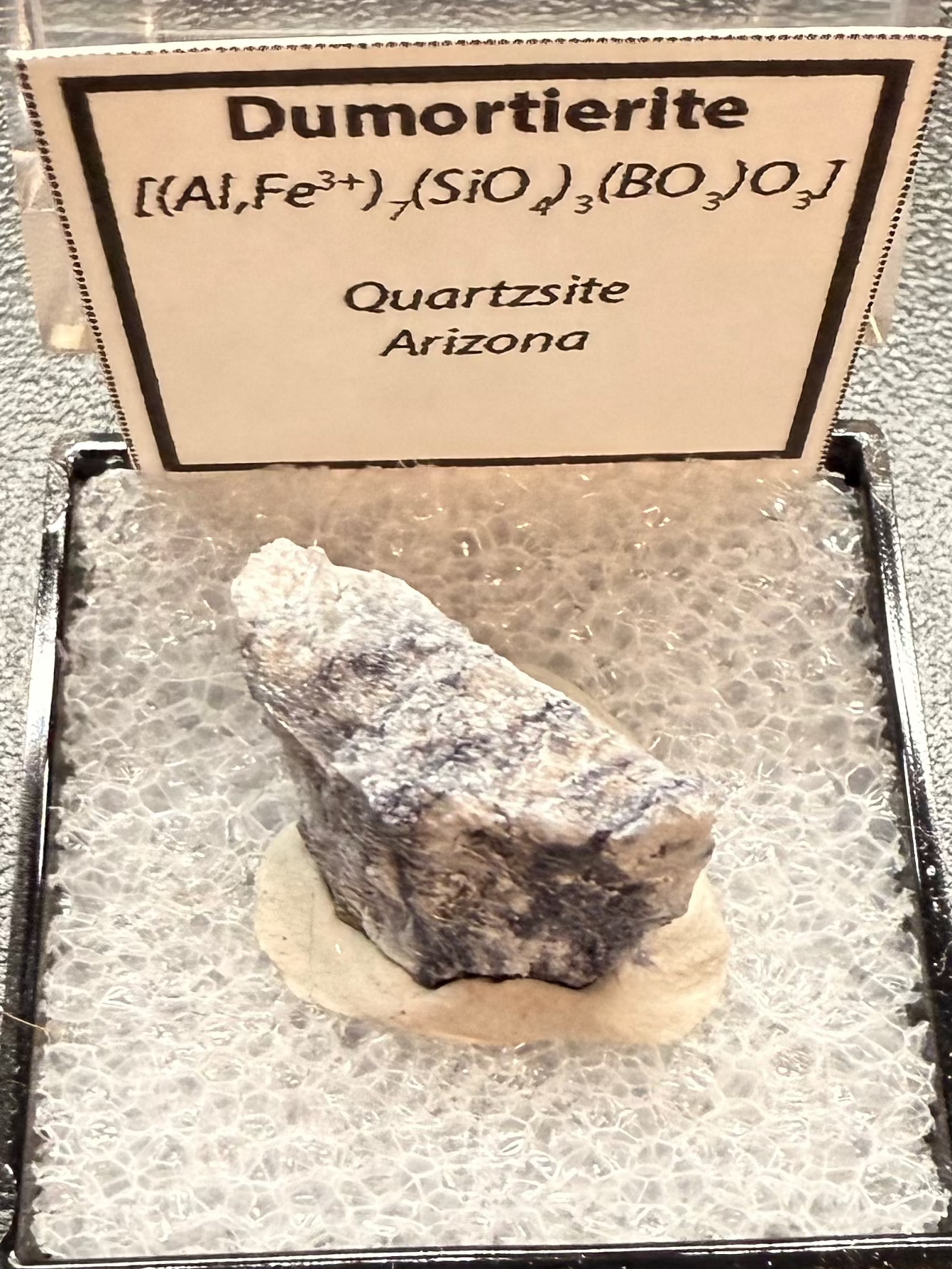 Dumortierite mineral in a display case with a label on a dark surface