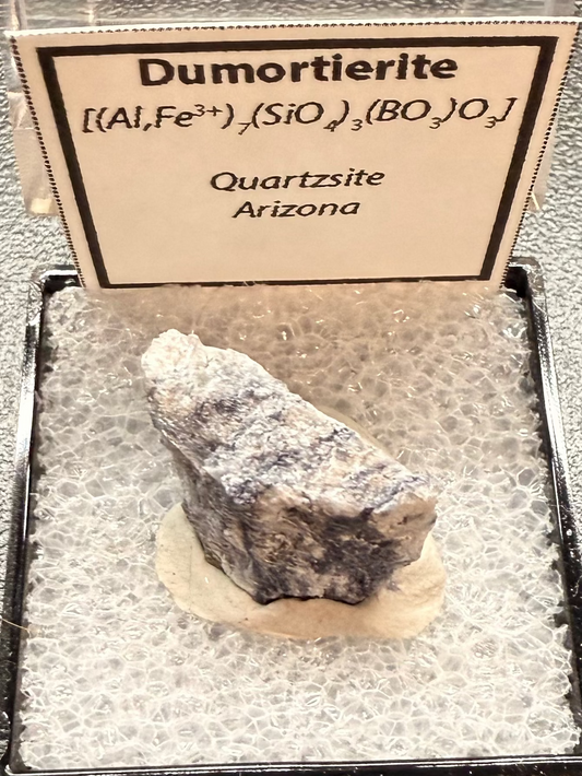 Dumortierite mineral in a display case with a label on a dark surface