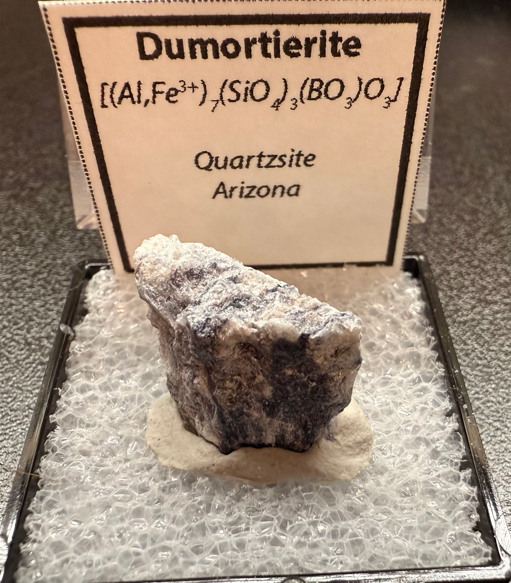 Mineral sample in a display case with a label on a dark surface