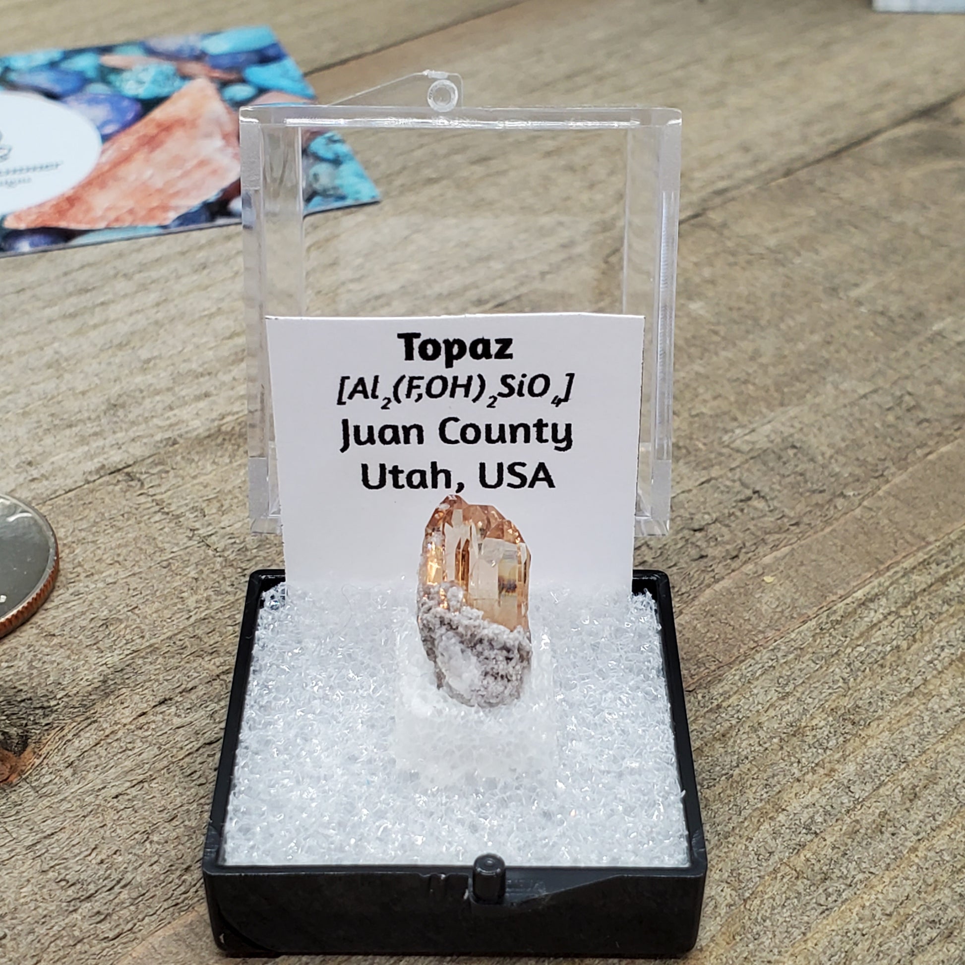 Peach colored topaz crystal in box with formula and origin label.
