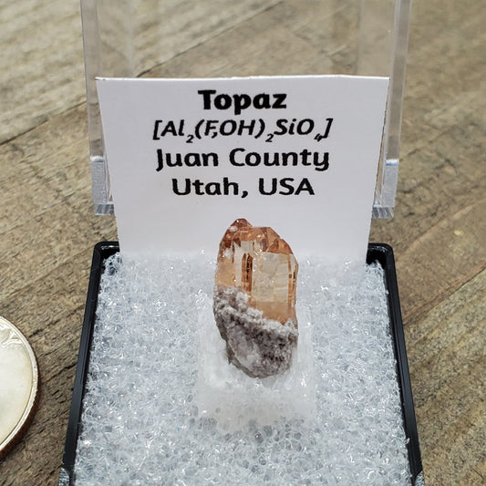 Peach colored topaz crystal in box with formula and origin label.